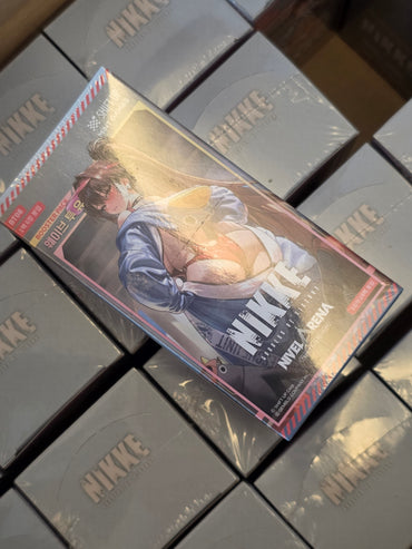 A boxed product labeled NIKKE on the cover, photographed at an angle on top of multiple stacked gray NIKKE boxes. The front box features a stylized illustrated figure in blue clothing with a large censored blur block over the center.