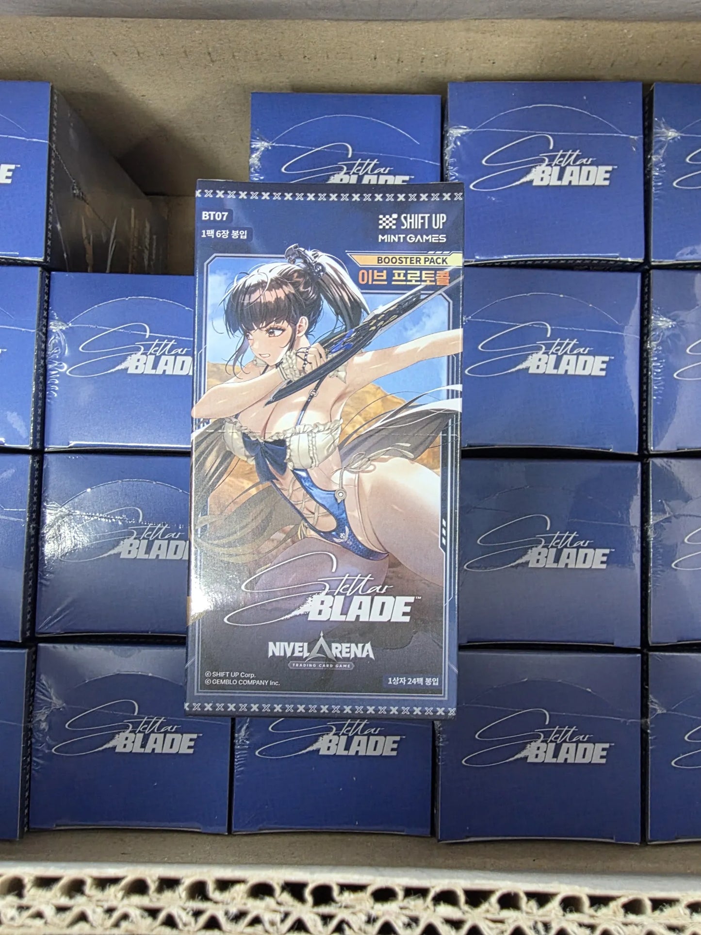 Sealed Nivel Arena Stella Blade booster boxes stacked on top of other boxes inside a cardboard box. The box feature game artwork of a female character , eve with long brown hair against a dark blue background.