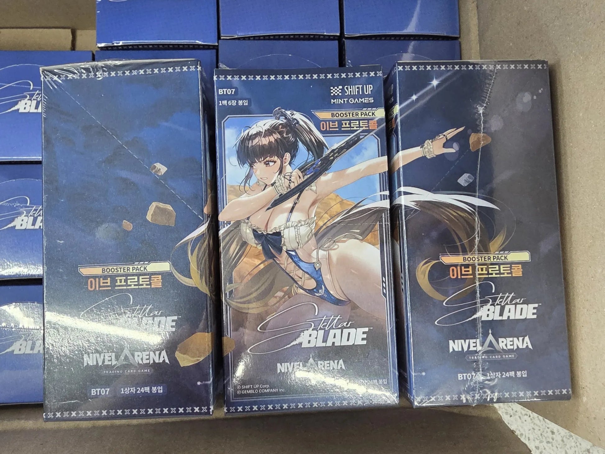 Three sealed Nivel Arena Stella Blade booster boxes stacked on top of other boxes inside a cardboard box. The box feature game artwork of a female character , eve with long brown hair against a dark blue background.